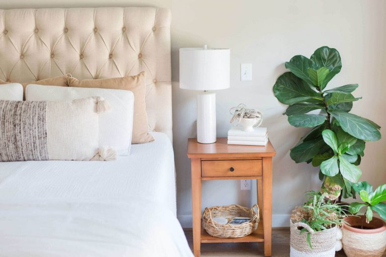 cozy bed with high, buttoned headboard, wooden side table with a white lamp next to leafy plant. The natural light coming in gives this bedroom a light, cozy and airy feel to the bedroom.